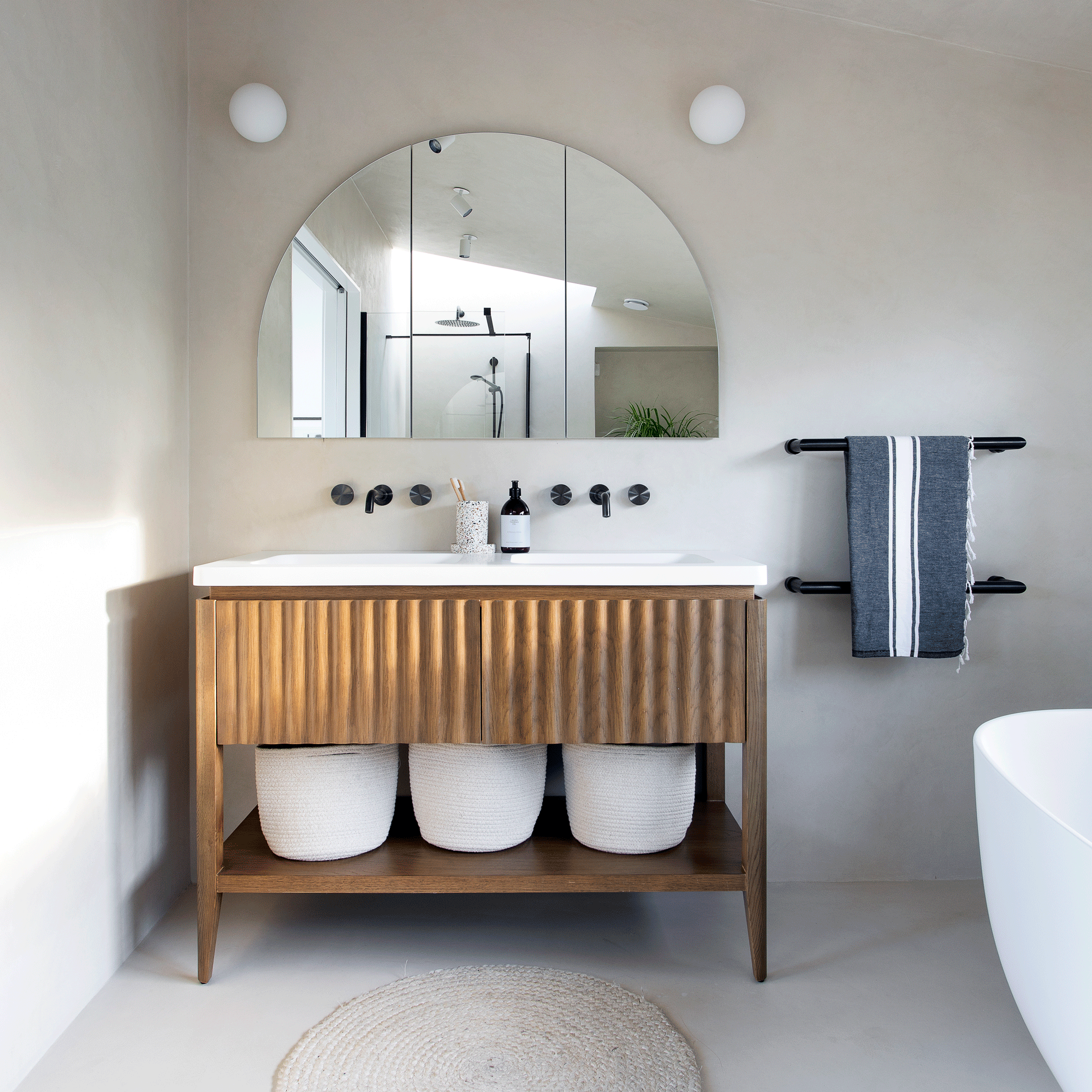 a bathroom with a wooden fluted vanity unit with white twin basins and black wall hung tap and a half moon mirror above