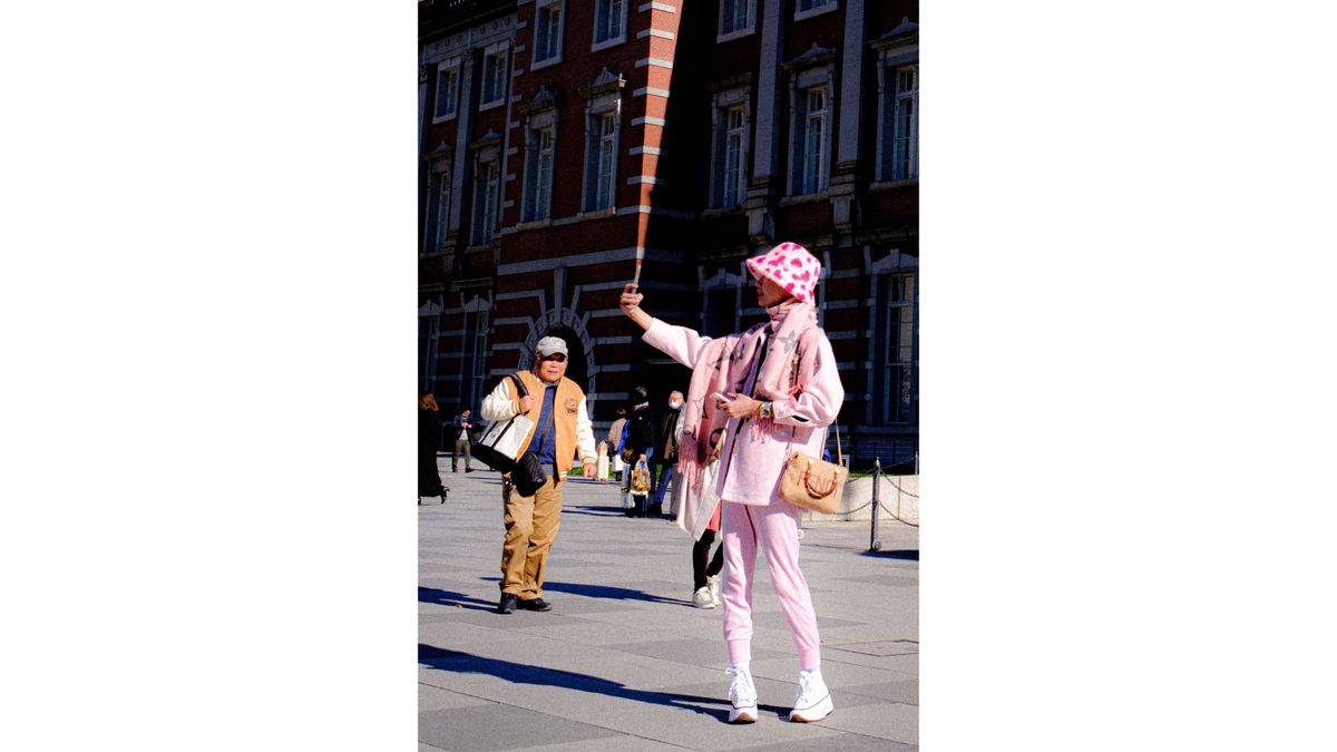 A person in pink clothing and a heart-patterned hat takes a selfie outside a large brick building, while another person stands in the background