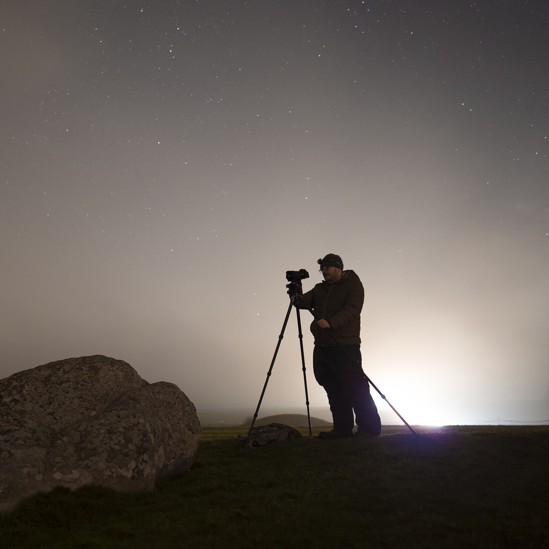 A silhouette of a person photographing the night sky with a tripod, surrounded by stars and a soft glow in the background