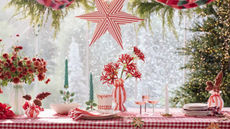 Festive tablescape featuring a red plaid tablecloth, candy-cane striped ceramic tableware and pendant lights, twisted evergreen taper candles, and warm string lights softly aglow in the background.