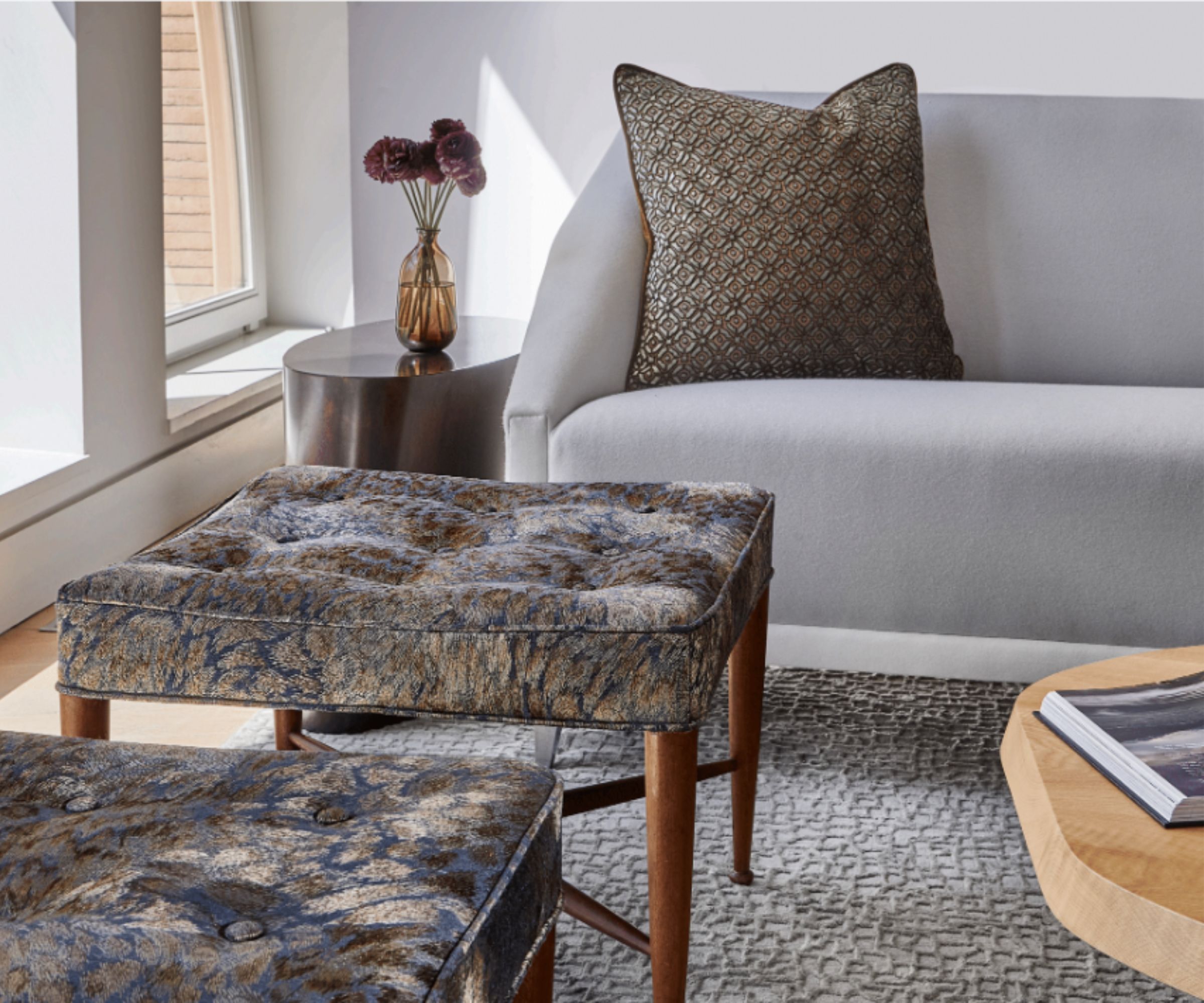 Close up of cream sofa with brown patterned cushion, textured cream rug, footstool with wooden frame and brown upholstery, metal side table with vase in background
