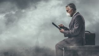 A smartly dressed man sat in a polluted city wearing a facemask while using a tablet computer