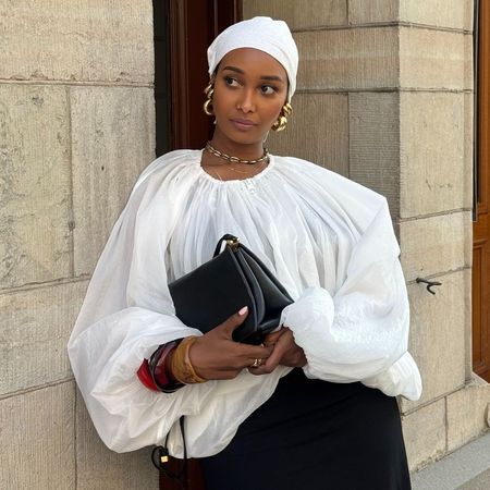 Woman wears a white bandana, white blouse, gold jewellery and black trousers