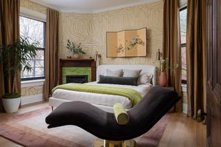 A bedroom with a patterned wallpaper, a curving laidback chair and a white bed