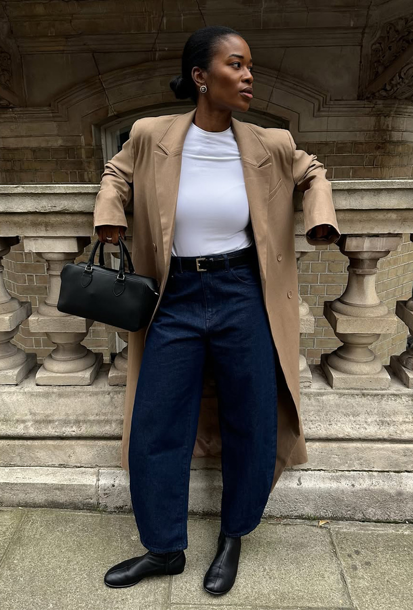 A woman standing outside wearing a long brown wool coat over a white T-shirt styled with dark-wash barrel-leg jeans, black flat ankle boots, a black bowler bag, and silver earrings.