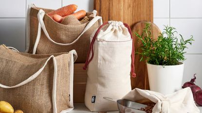 A set of cotton and jute food storage bags on a kitchen counter, beside some wooden cutting boards and a metal plant pot of herbs. 