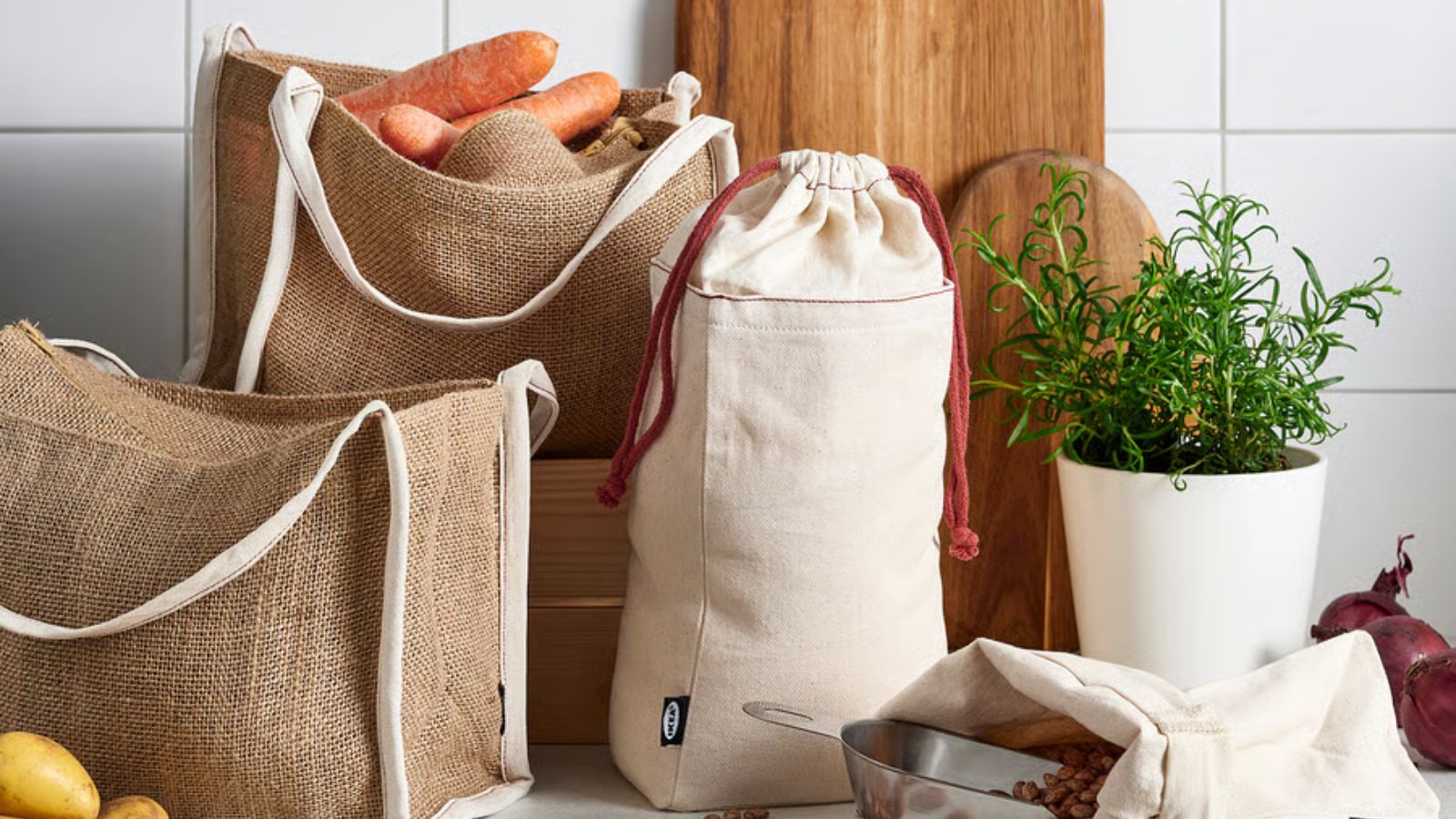 A set of cotton and jute food storage bags on a kitchen counter, beside some wooden cutting boards and a metal plant pot of herbs. 
