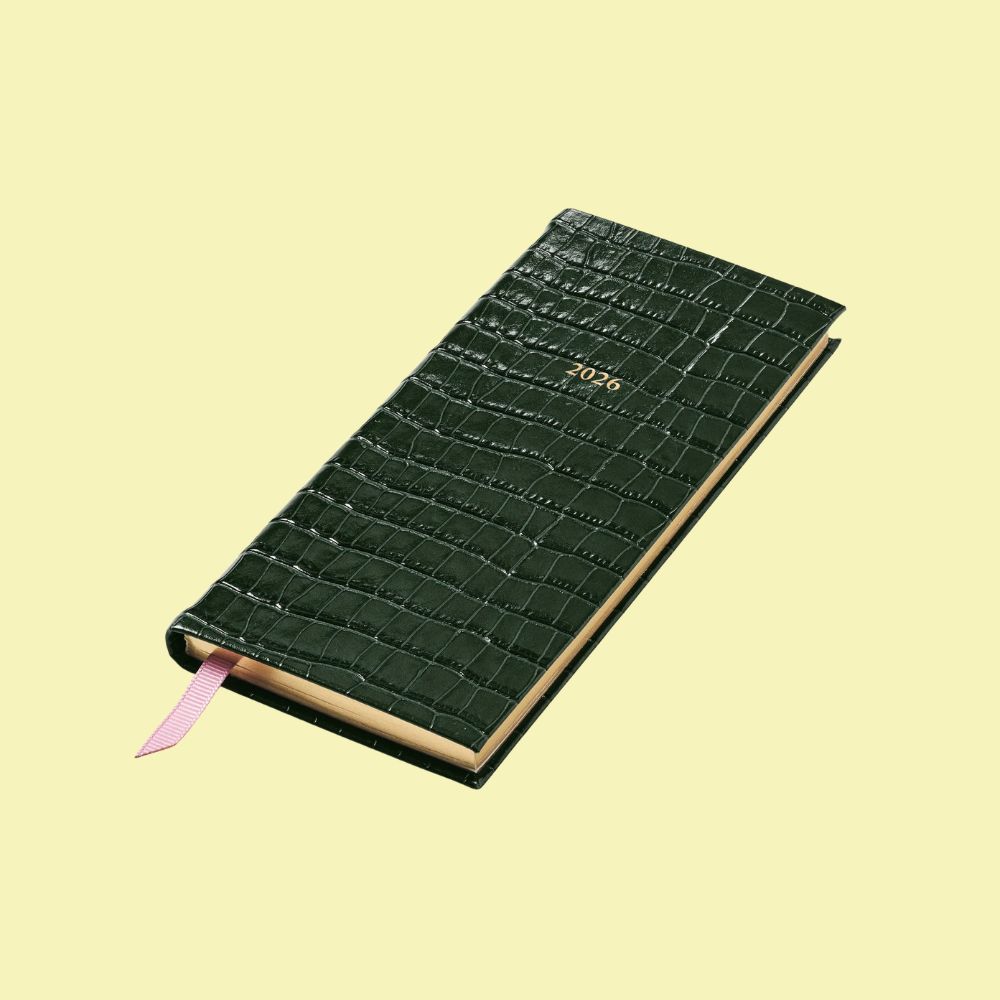 A pocket-size, slim leather planner in emerald green with a gold detail "2026" inscription on it sits sideways against a pale yellow background.