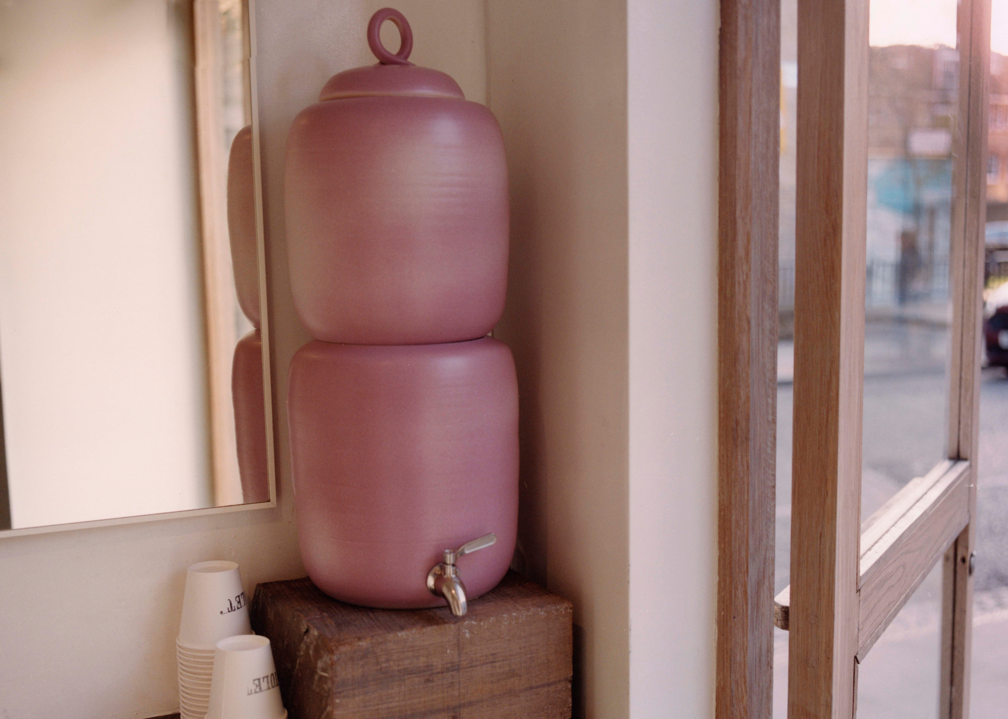 A dusty pink ceramic water vessel on a wood block near a mirror and stacks of paper cups by a wood and glass door