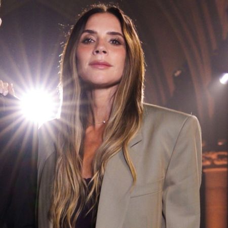 Victoria Beckham poses at the Victoria Beckham SS27 runway show with David Beckham. She wears a grey suit with brown pointed-toe heels.