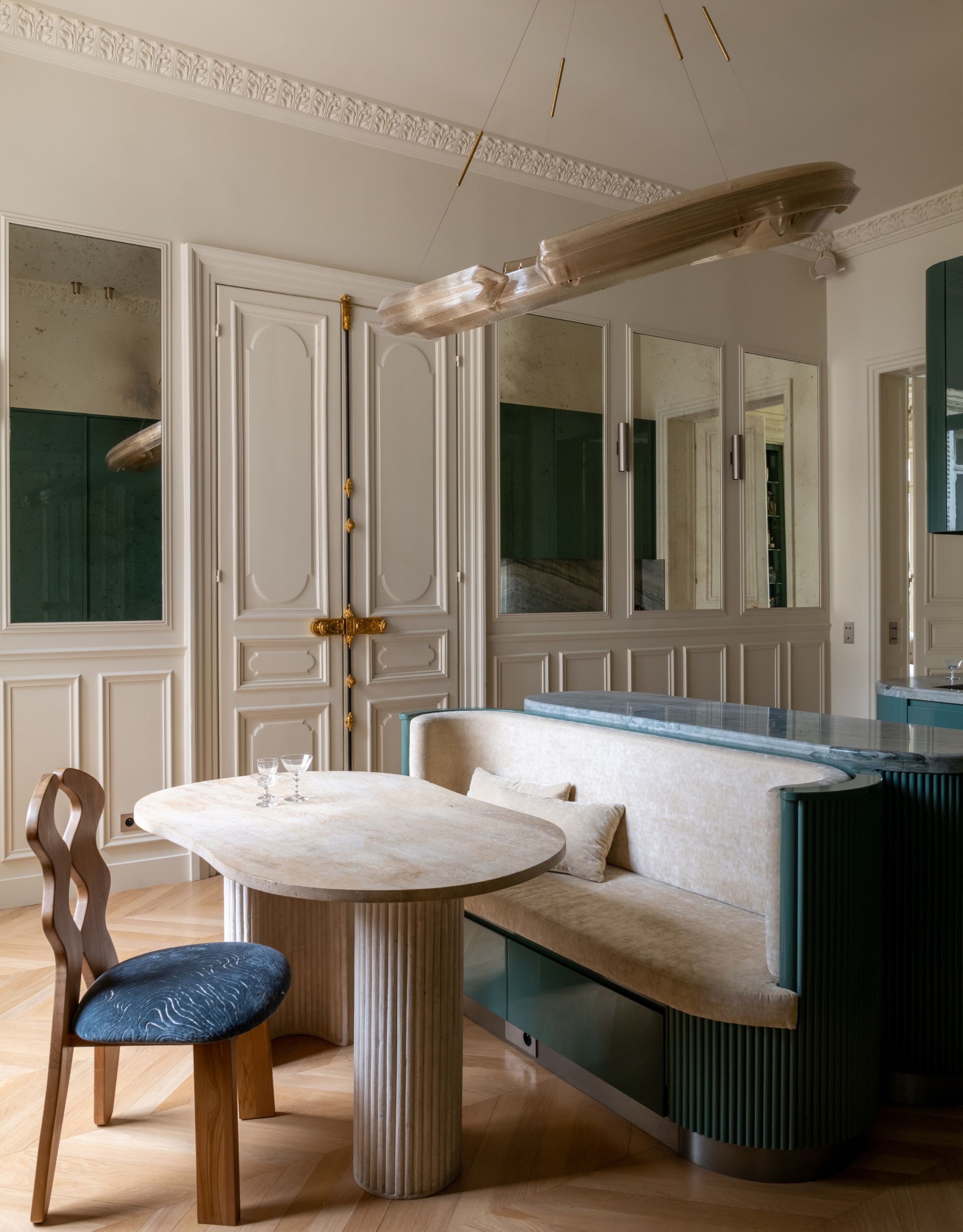 A grand dining room with off-white walls, architectural moldings and paneling, including mirror panels, with an off-center circular pendant light above a banquette and table