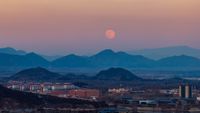 A full moon is pictured rising in the evening sky over a serene, misty mountain range bordering a city.