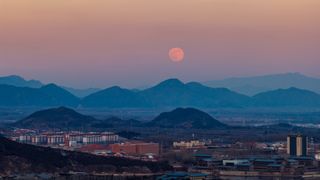 A full moon is pictured rising in the evening sky over a serene, misty mountain range bordering a city.