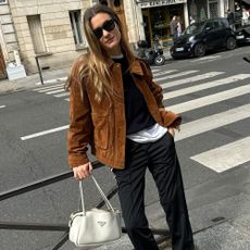French influencer @annelauremais stands outside in Paris wear a brown suede jacket with black trousers and transparent heels.