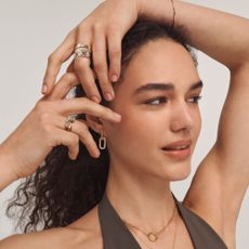 Woman posing with silver and gold rings on her fingers, a gold necklace, bracelet & earrings. In front of a plain background