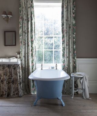 Bathroom curtain ideas with floral curtains framing a garden view, with blue freestanding bathtub