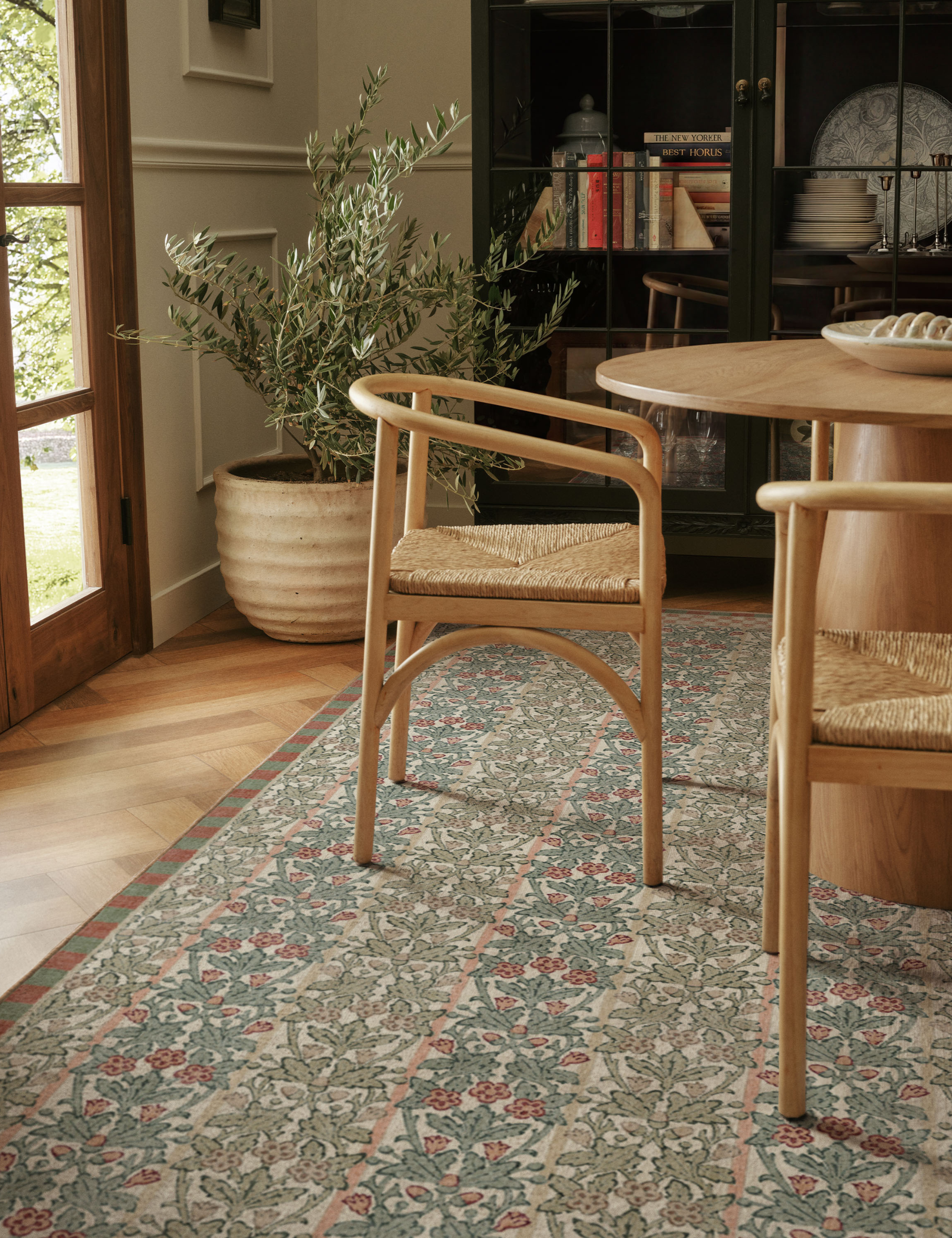 Neutral minimalist dining room vignette featuring mid-century-inspired rattan dining chairs, a wooden pedestal table, an oversized potted plant, and a botanical blue-and-green area rug.