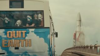a large rocket looms in the distance behind a bus with the words "Quit Earth" on it