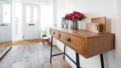 Tidy white entryway with wooden console table with metal legs, vase with flowers, two metal wire candle holders with burning candles, letter holder rack. Hardwood oak flooring, mirrored storage cabinet and white front door