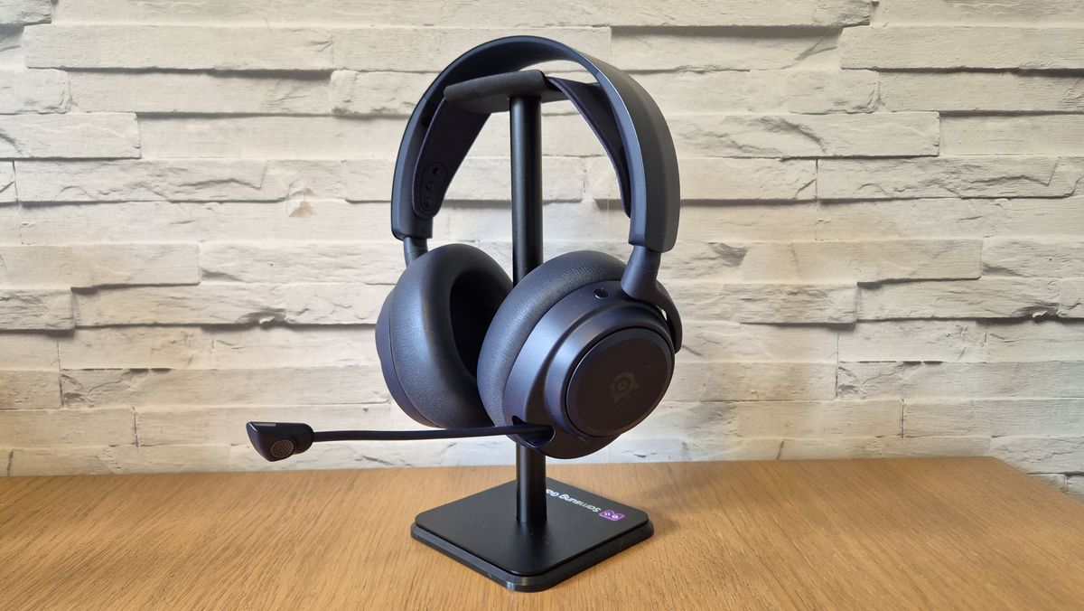 The SteelSeries Arctis Nova Pro Omni headset sitting on a headset stand in front of a white brick background and on a wooden surface.