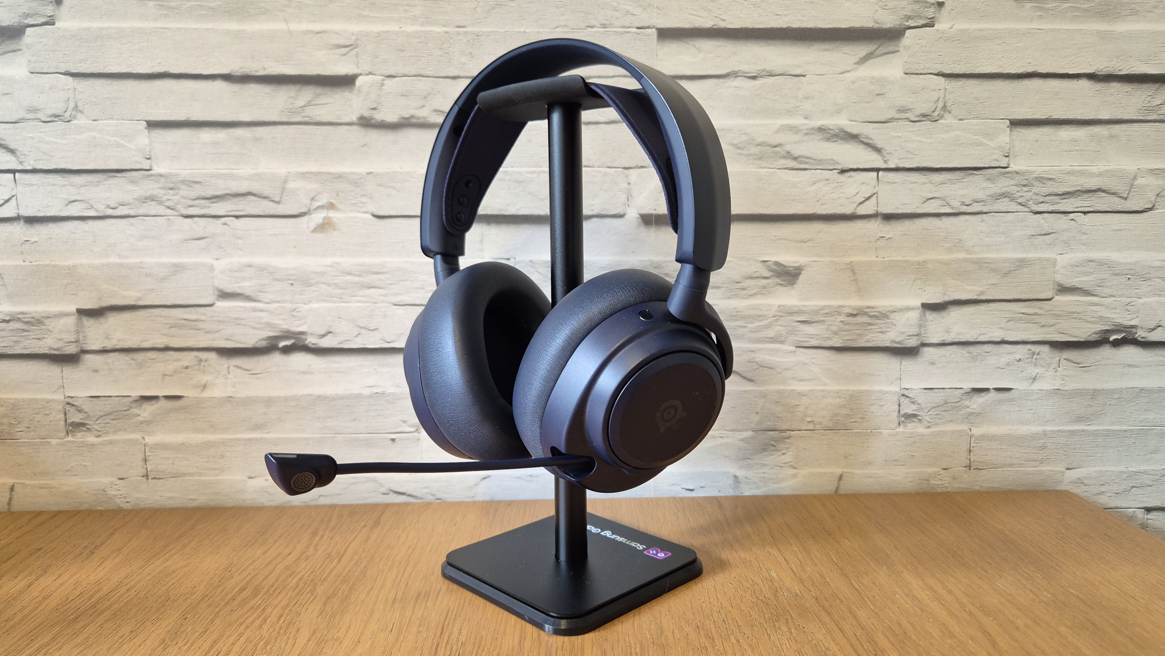 The SteelSeries Arctis Nova Pro Omni headset sitting on a headset stand in front of a white brick background and on a wooden surface.