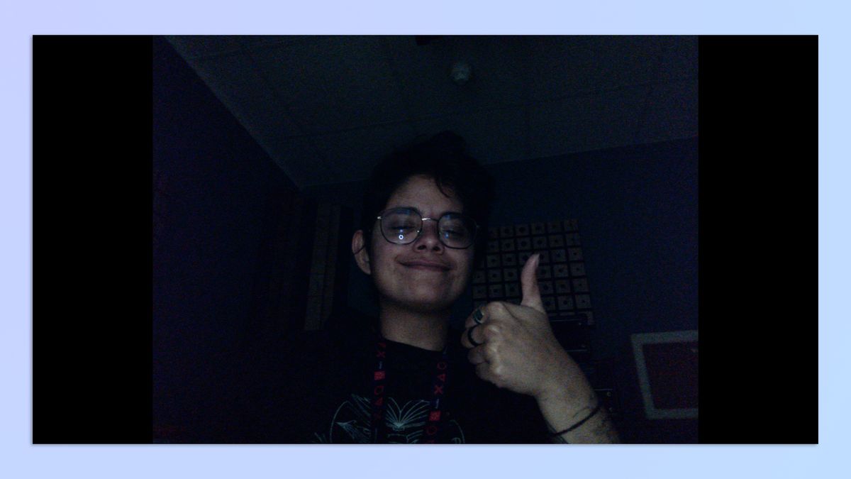 A woman with short hair sitting in a dark room, holding up her hand in the shape of a thumbs-up