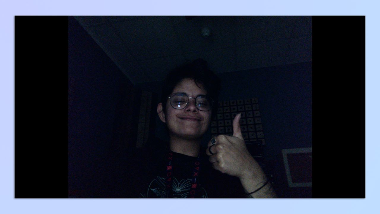 A woman with short hair sitting in a dark room, holding up her hand in the shape of a thumbs-up