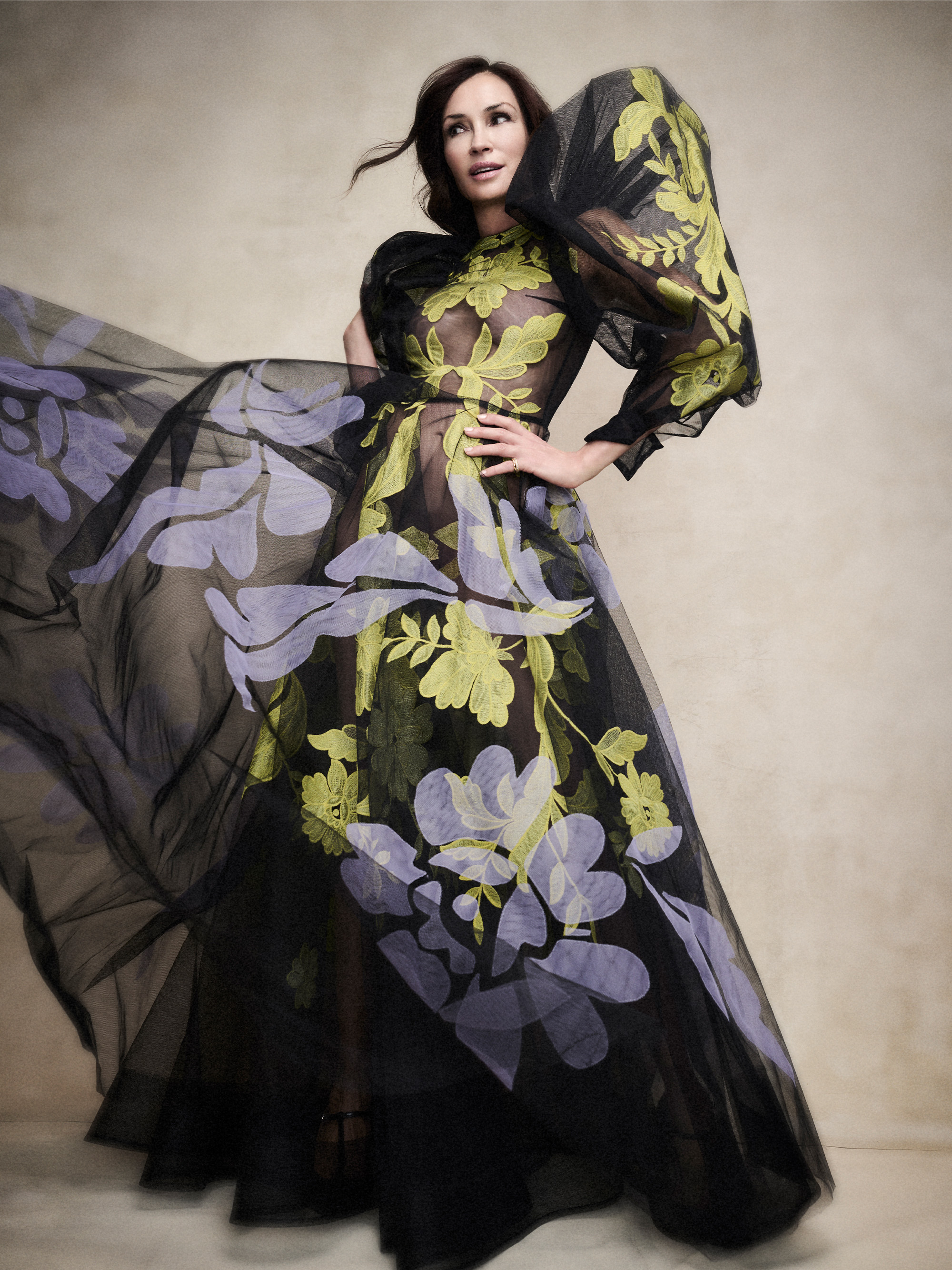 Photo of Famke Janssen posing in front of a tan seamless backdrop. She is wearing a black sheer dress with yellow and purple floral embroidered and oversized sleeves.