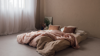 A bed with dusty pink bedding on the floor of a bedroom with the sun shining through the window