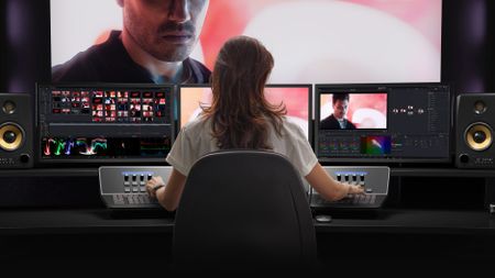 A person sitting in an office chair is using two specialized control panels to edit video across a three-monitor setup, flanked by studio monitor speakers and with a large screen showing a close-up of a man above.
