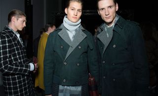 Four male models wearing looks from Canali's collection. They are wearing light coloured shirts and jumpers. One model is wearing a yellow coat and another is wearing a black and white coat with grid style pattern and holding a pair of gloves. The remaining two models are wearing teal jackets and coats with one of them holding a red bag