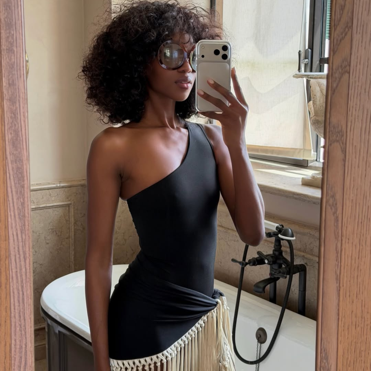 Emmanuelle wearing a black one-shoulder swimsuit with a tassel cover-up