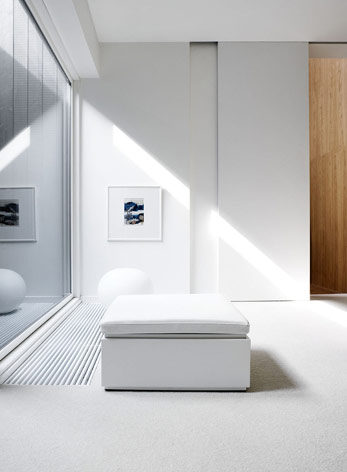 The master bedroom has a white carpet and white walls. On the wall, there is a photo set in a white frame, with a white tabouret.