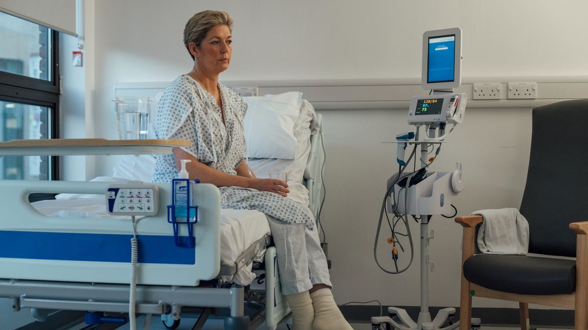 Middle-aged woman is pictured sitting on a hospital bed wearing a surgical gown. There is hospital equipment in the background. Her legs are crossed and she is looking in front of her with a concerned look on her face. There is a chair to the right of her.