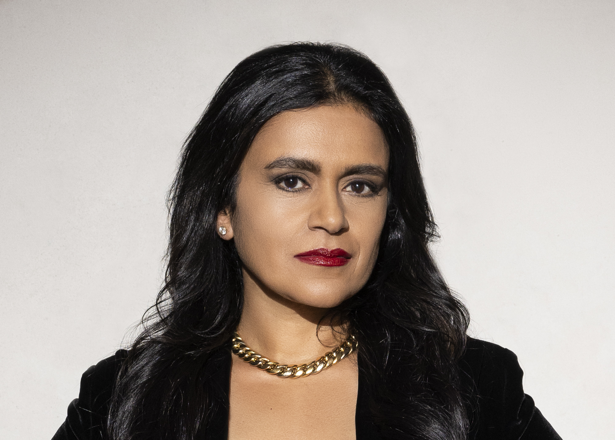 A headshot of a woman wearing a black jacket and gold necklace, standing against a wall