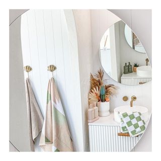 Brass ginko shell design towel hools on wall with chalky pastel patterned towels, round wall mirror, brass tap, round sink, and dried flower arrangement on vanity counter.