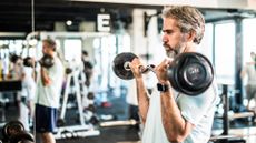 man standing next to a mirror in a gym curling a small weights bar up to his biceps, standing side on to the camera.