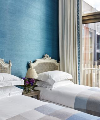A twin bedroom with large window, blue grasscloth wallpaper and white French-style rattan beds.