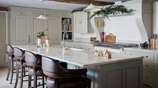 A festively decorated kitchen with white cabinets, white backsplash and range hood, and a large kitchen island with white marble countertop and four leather barstools. 