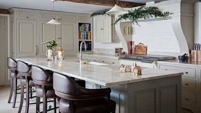 A festively decorated kitchen with white cabinets, white backsplash and range hood, and a large kitchen island with white marble countertop and four leather barstools. 