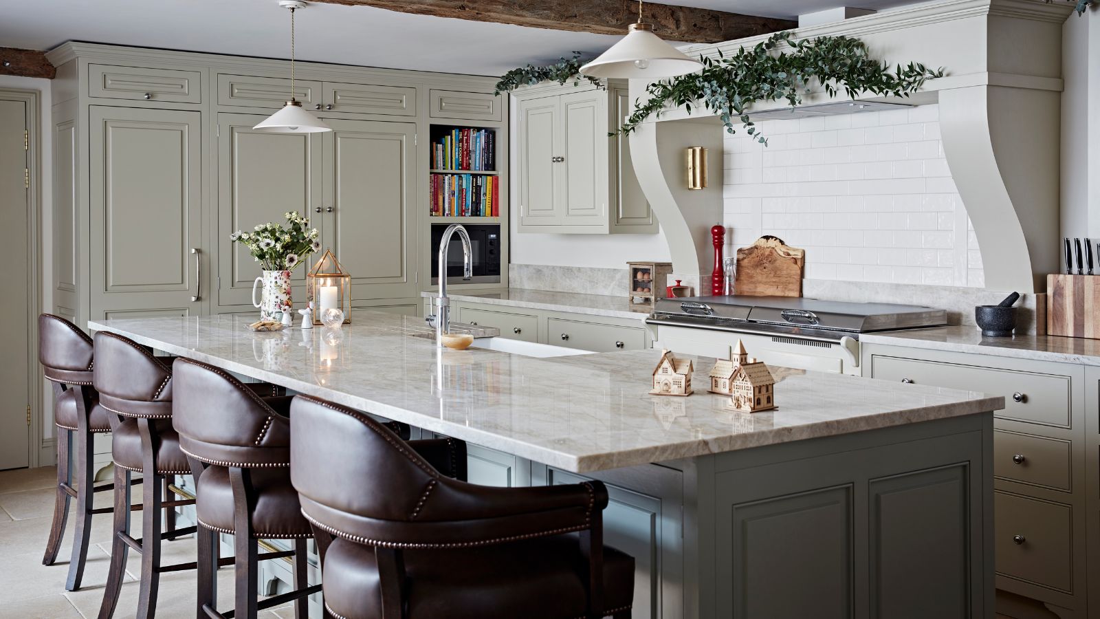 A festively decorated kitchen with white cabinets, white backsplash and range hood, and a large kitchen island with white marble countertop and four leather barstools. 