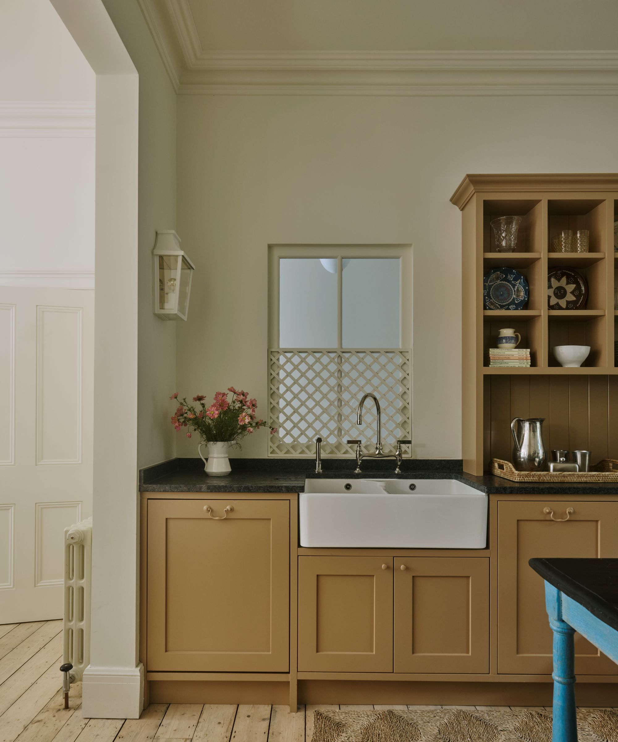 Farmhouse kitchen with caramel-coloured cabinetry, a white ceramic sink, a blue wooden island table, wooden floorboards, and open shelves with ceramics.