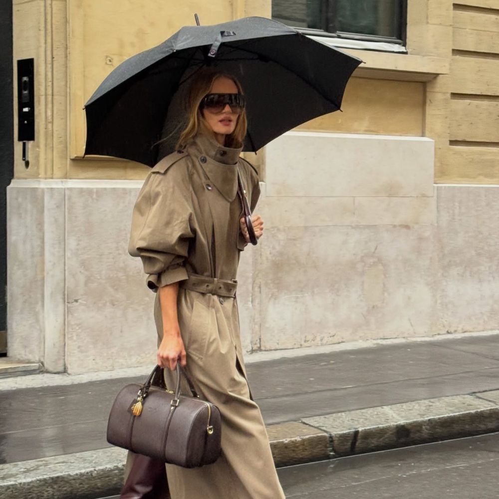 Model Rosie Huntington-Whiteley walks outside in the rain underneath an umbrella, wearing a beige funnel-neck trench coat with burgundy boots and a bowling bag.