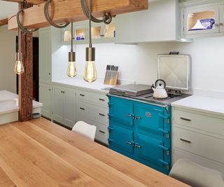 three exposed bulbs on long light cord wrapped around ceiling beam above wooden kitchen island, run of pale green kitchen cabinets on back wall with turquoise range cooker