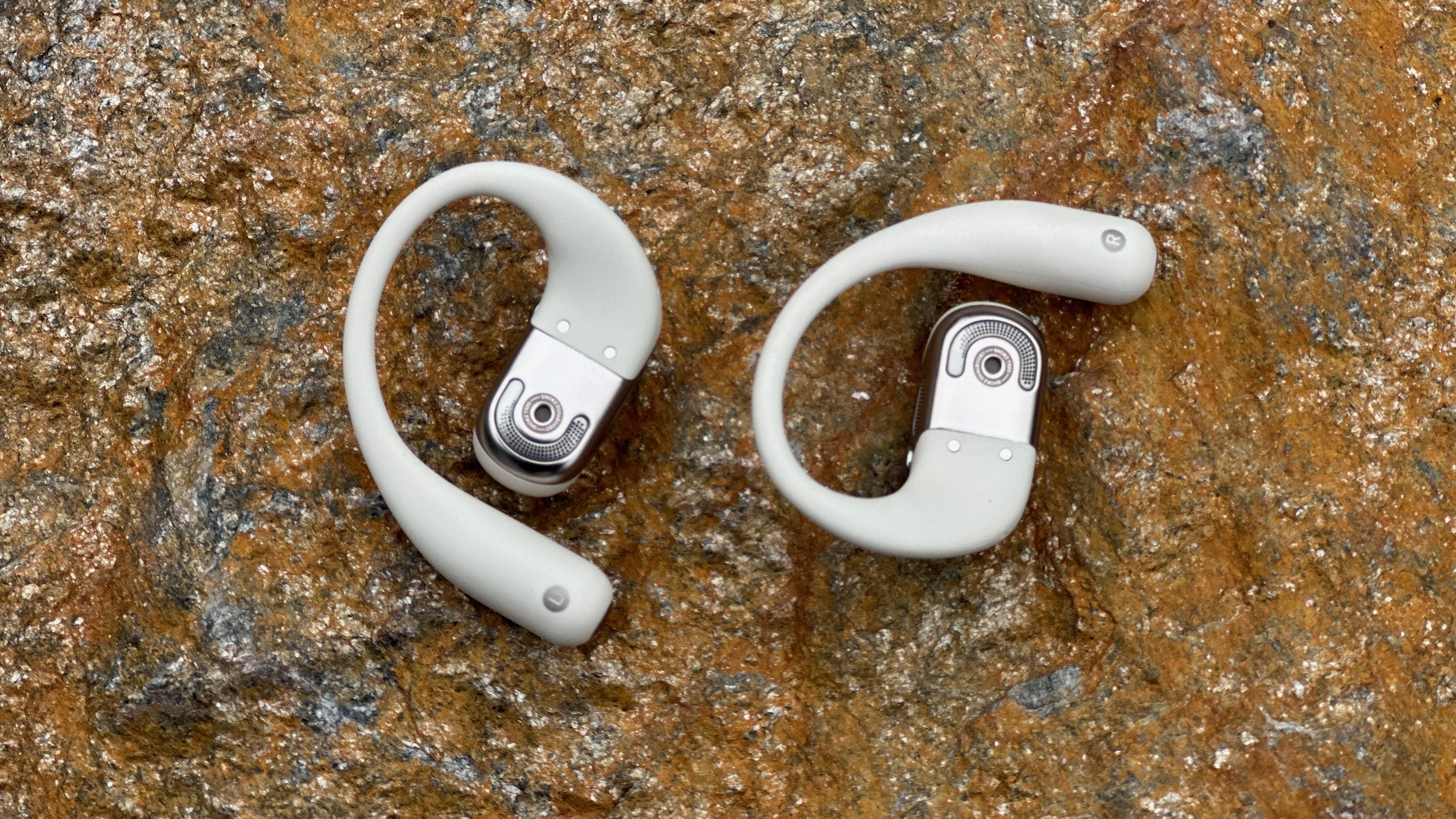 The Shokz OpenFit 2 earbuds sitting on a rock, both flipped over to show the main speakers that sit against your ears.