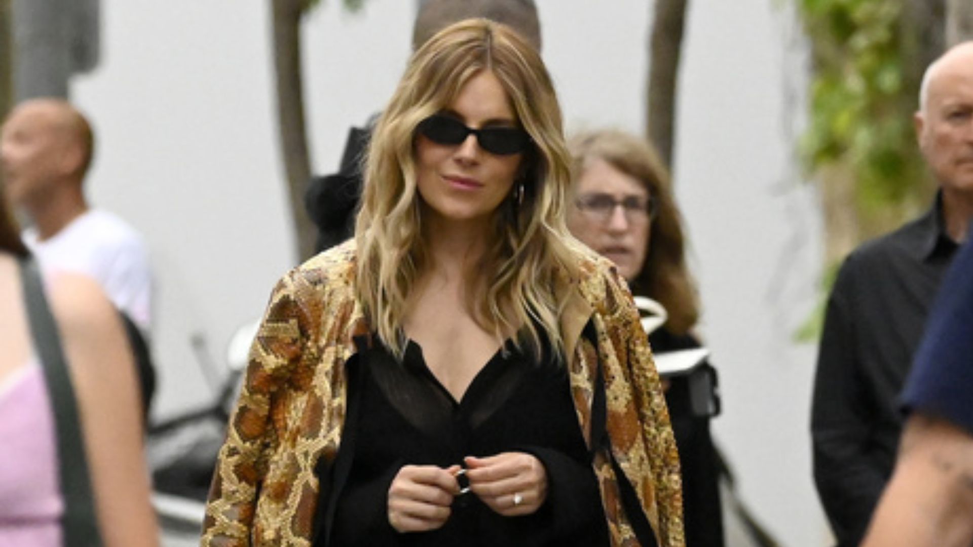 Sienna Miller walks down the street wearing blue baggy jeans with a snake print jacket and black strappy sandals.