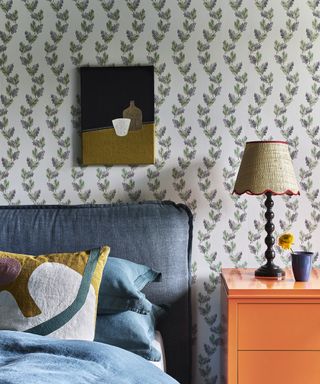 Relaxed and colourful bedroom with pretty leaf trail wallpaper design and a bright orange lacquer bedside cabinet