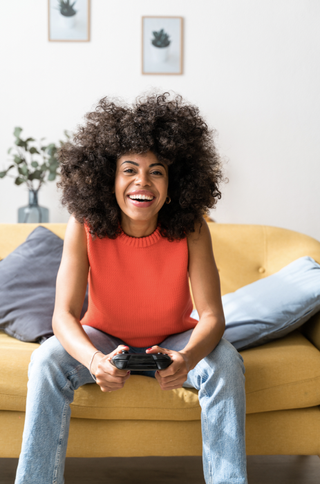 Optimistic woman sitting on living room sofa holding video game controller. Relaxed woman spending her free time at home, playing online with friends.