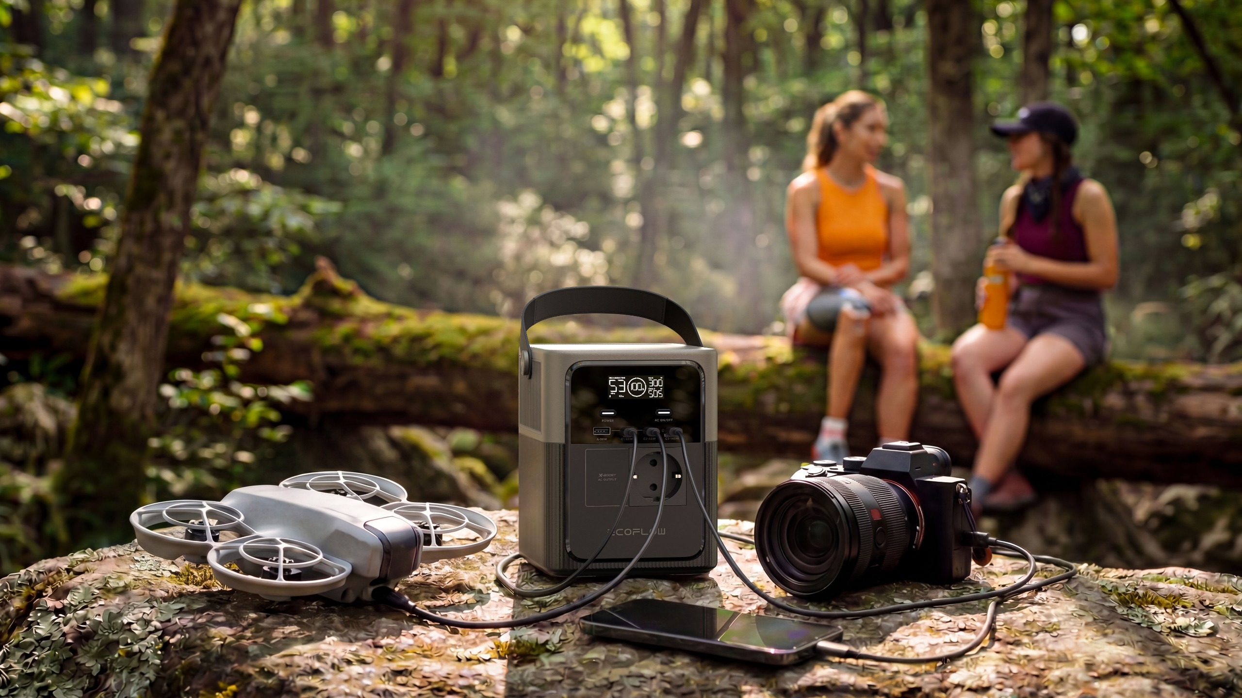 Set in a remotely wooded area, an EcoFlow portable power station charges a digital camera, smartphone, and drone in the foreground as two women hikers chat in the background.
