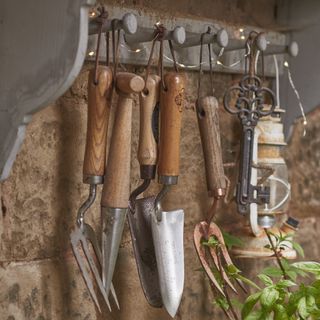 peg rail with garden tools hanging from it, stone wall behind